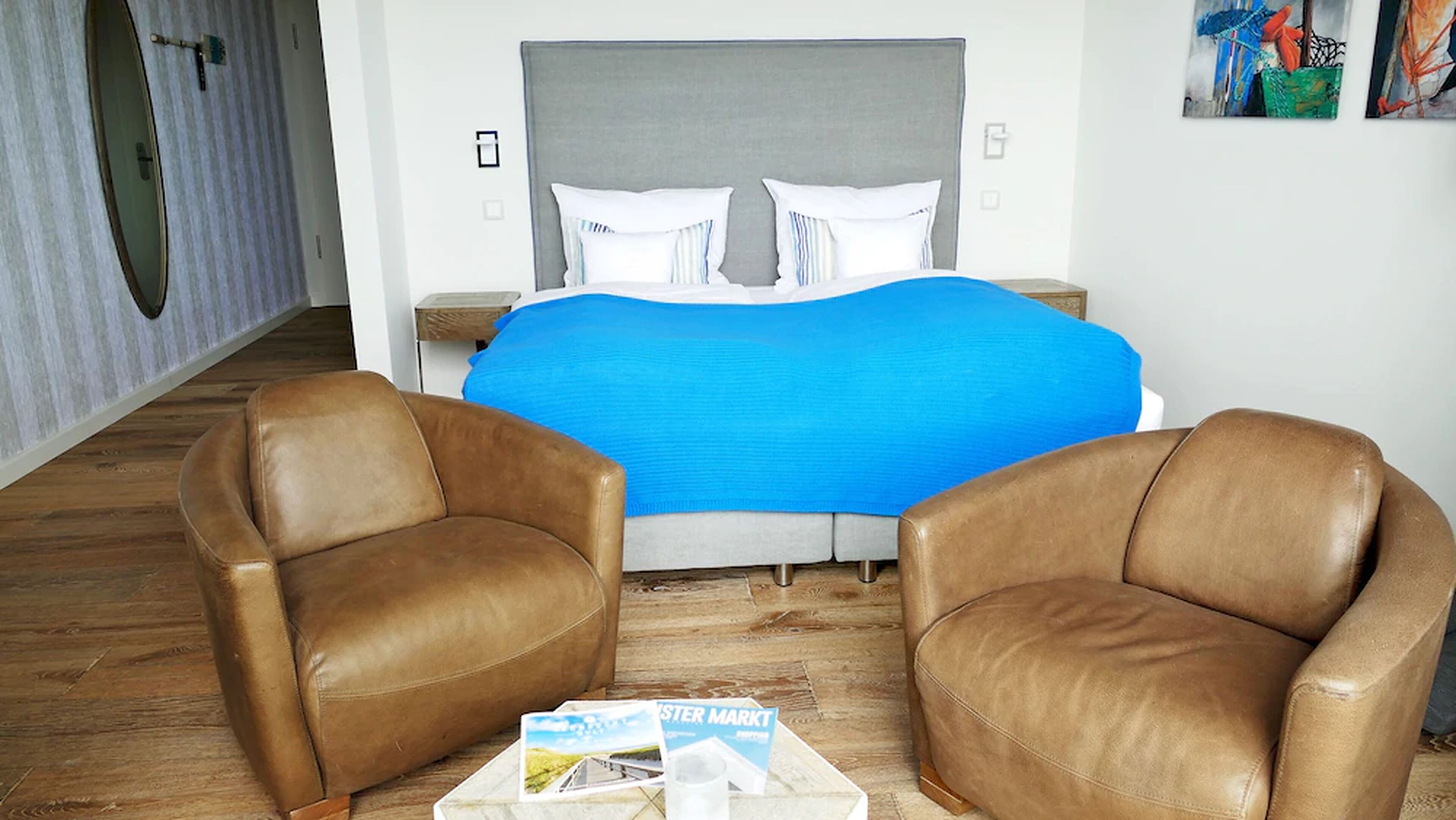 A hotel room with a bed, two leather armchairs, a nightstand, and a painting on the wall.