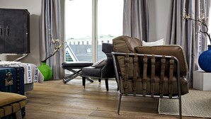 A modern living room with a leather sofa, a small table, and a cabinet. There is a window with curtains and a view of buildings outside. - Easy Living Hotel (List)