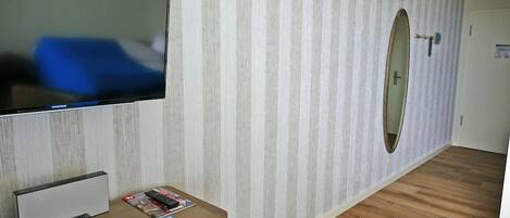 A hotel room with a flat-screen TV mounted on the wall, a wooden desk with magazines and a remote control, a mirror on the wall, and a wooden floor.