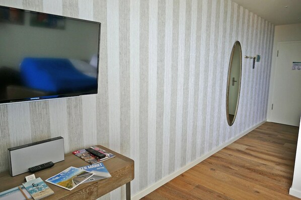 A hotel room with a flat-screen TV mounted on the wall, a wooden desk with magazines and a remote control, a mirror on the wall, and a wooden floor.