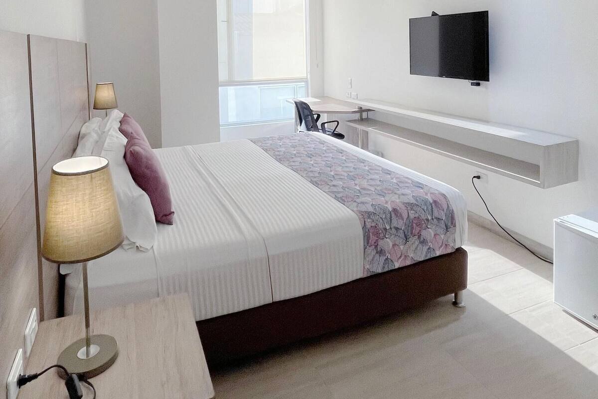 A modern hotel room with a bed, bedside table, lamp, TV, and a desk area.