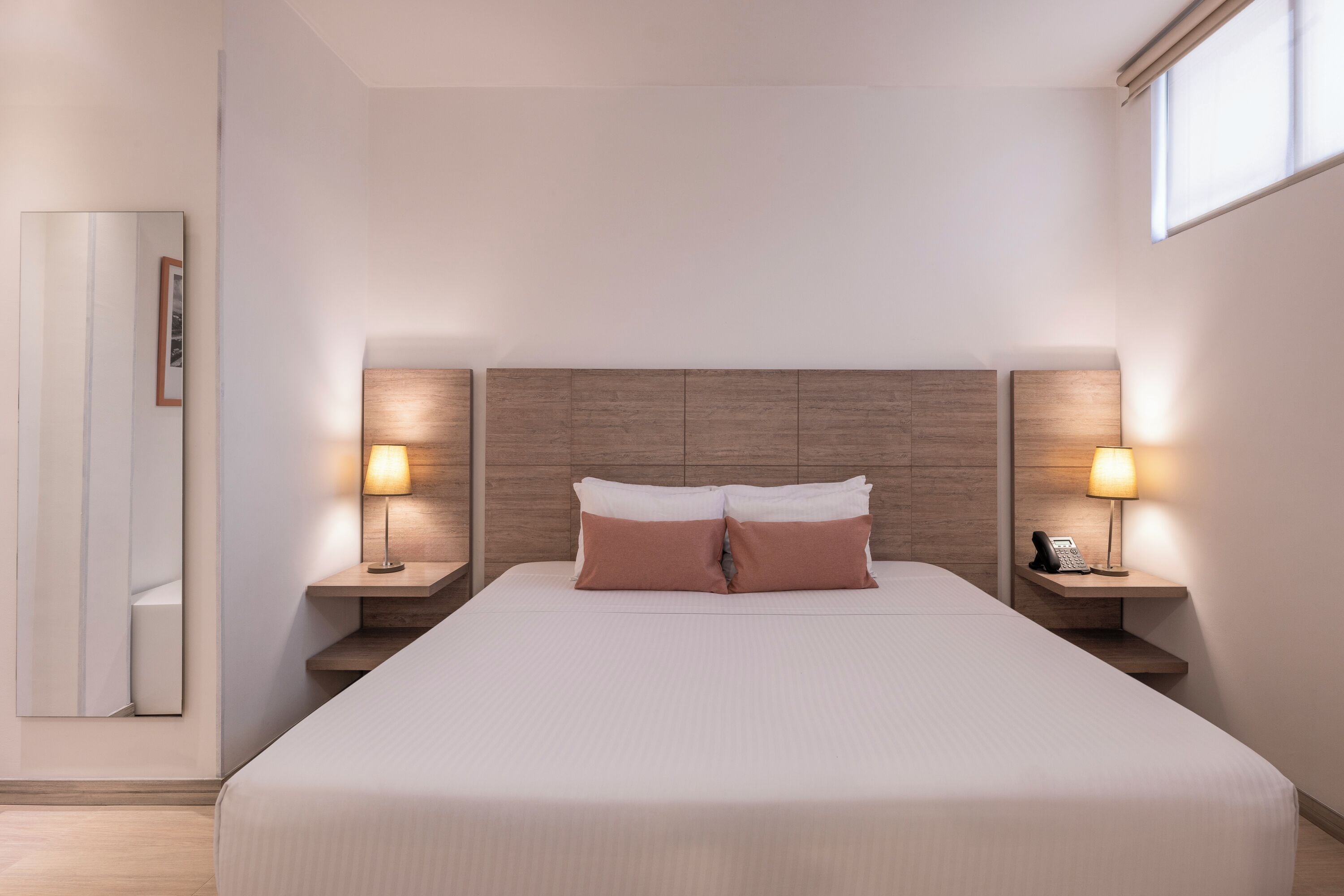Una habitación de hotel ordenada con una cama grande, dos mesitas de noche con lámparas, un espejo y un cuadro en la pared.