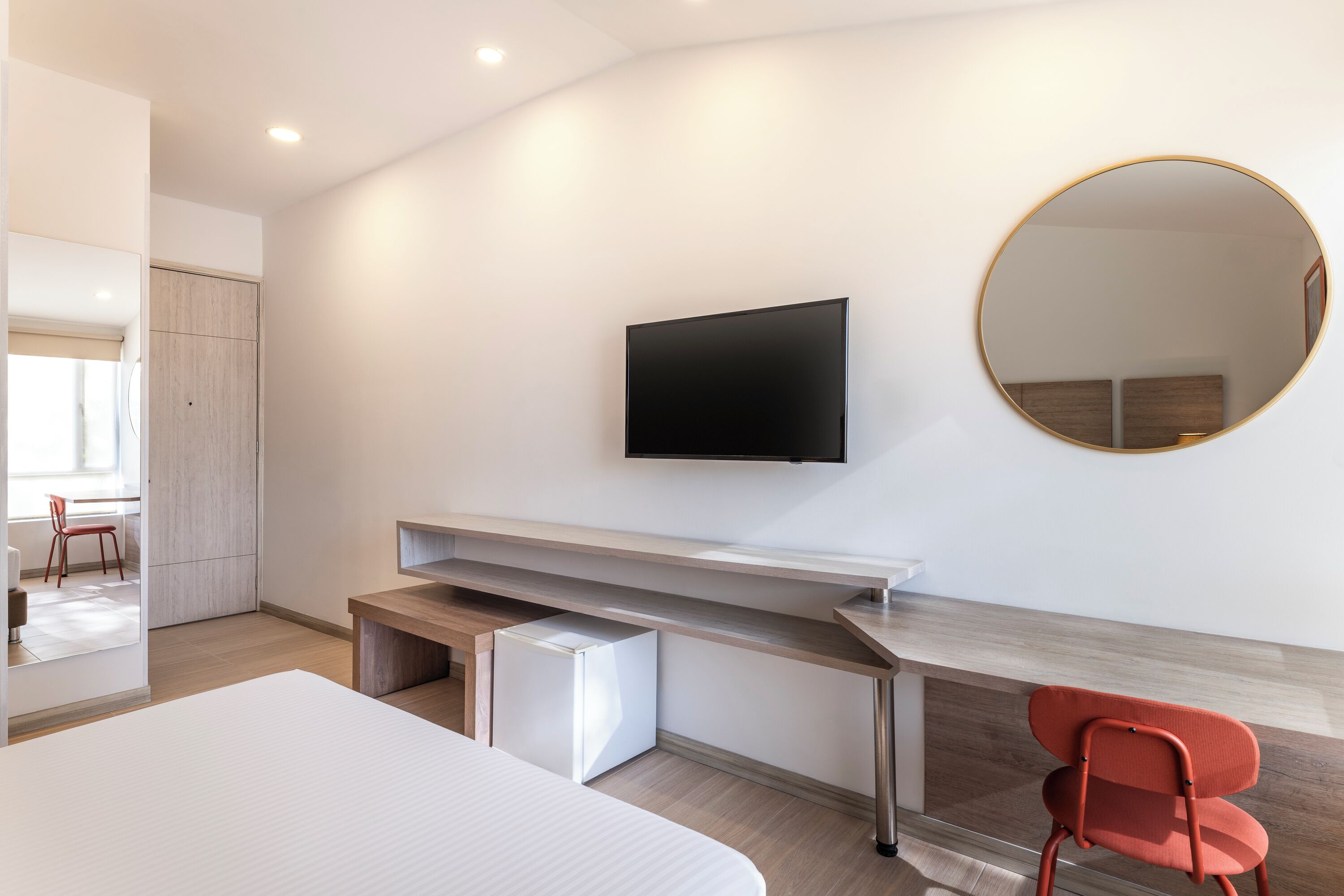 Une chambre d’hôtel moderne équipée d’un lit, d’un bureau avec un téléviseur à écran plat, d’un petit réfrigérateur et d’une chaise rouge.
