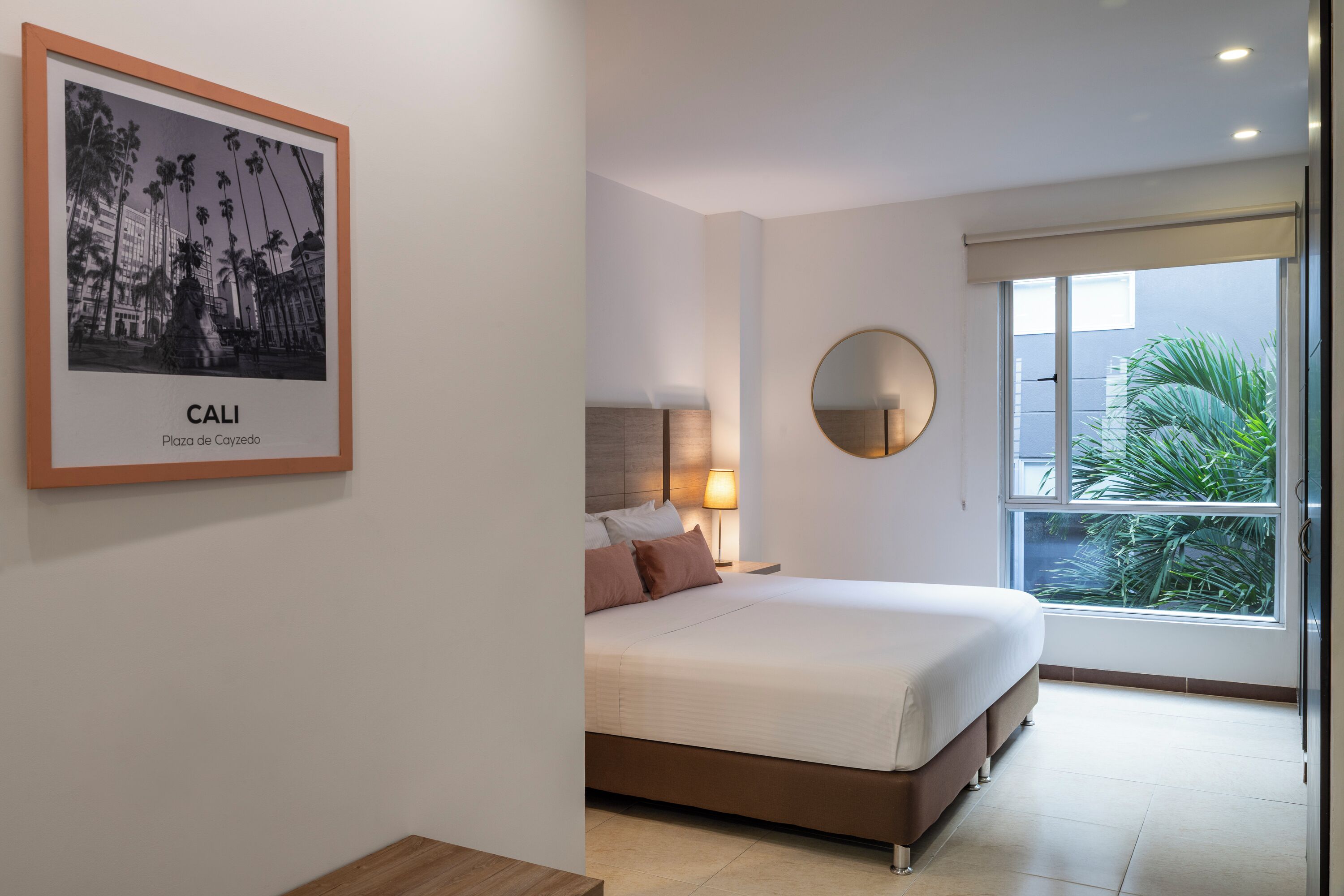 Un dormitorio moderno con una cama, un espejo redondo, una mesita de noche y un cuadro enmarcado de un paisaje urbano.