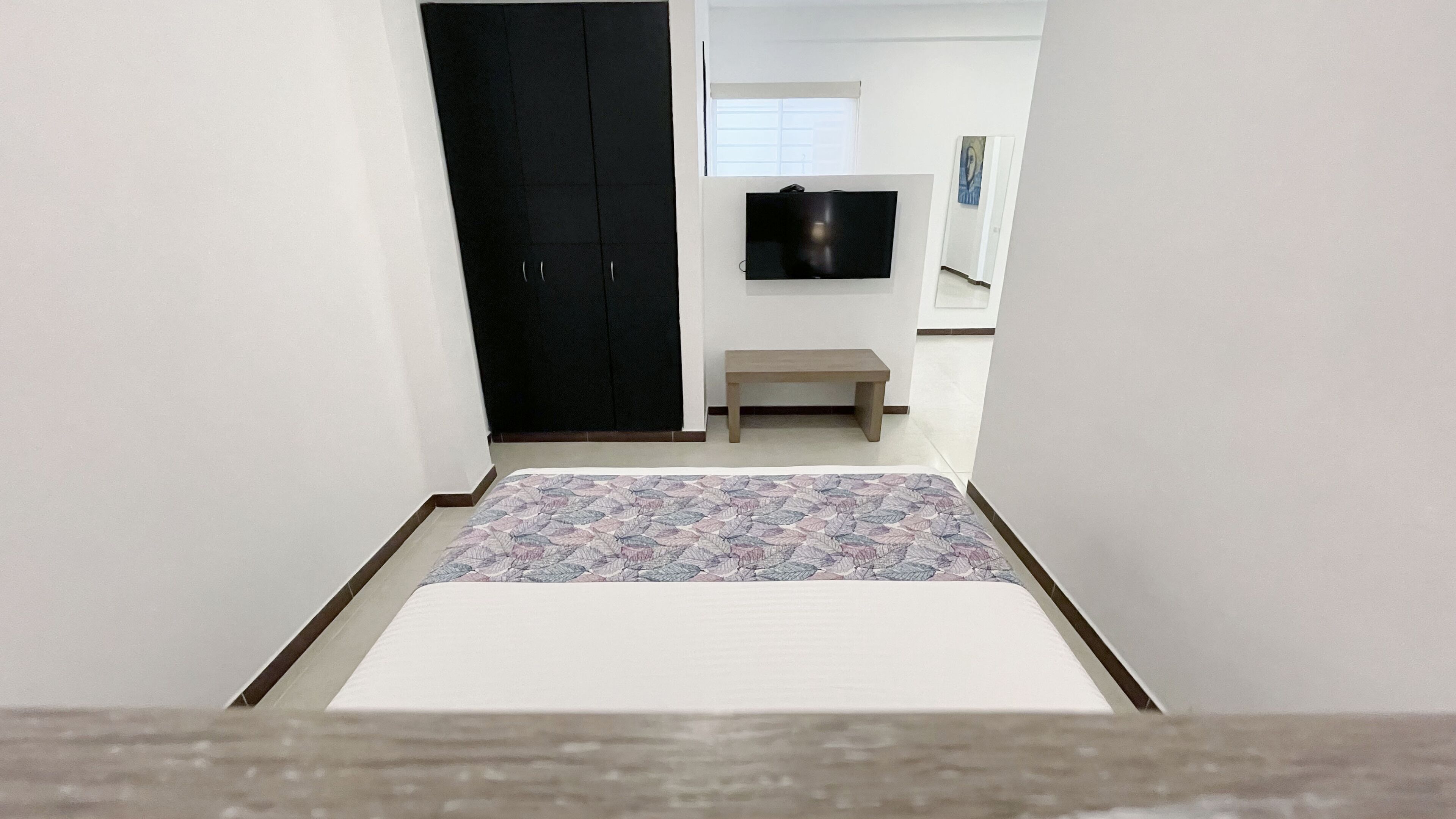 A bedroom with a bed, a dark wardrobe, a small bench, a television on the wall, and a patterned rug.