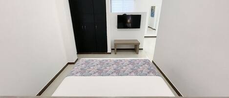 A bedroom with a bed, a dark wardrobe, a small bench, a television on the wall, and a patterned rug.