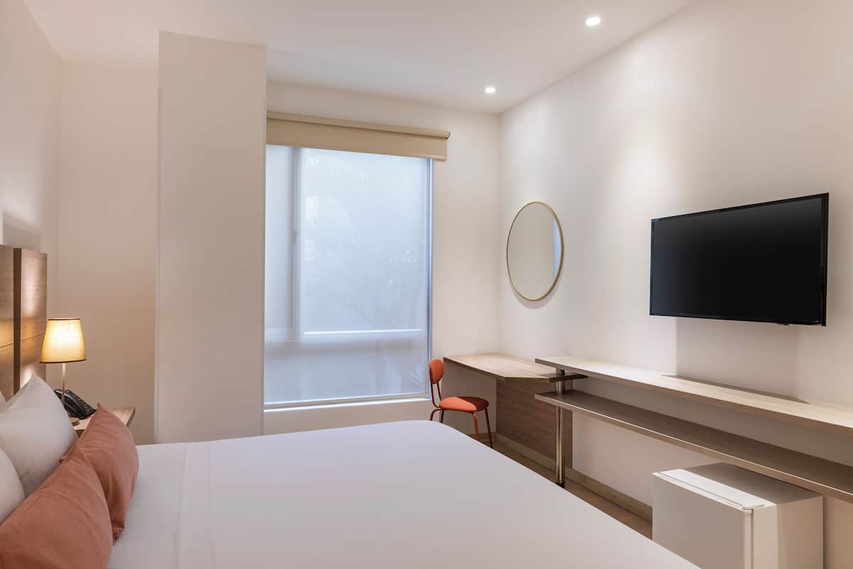 A modern hotel room with a large bed, a desk, a chair, a television, and a window with a view.