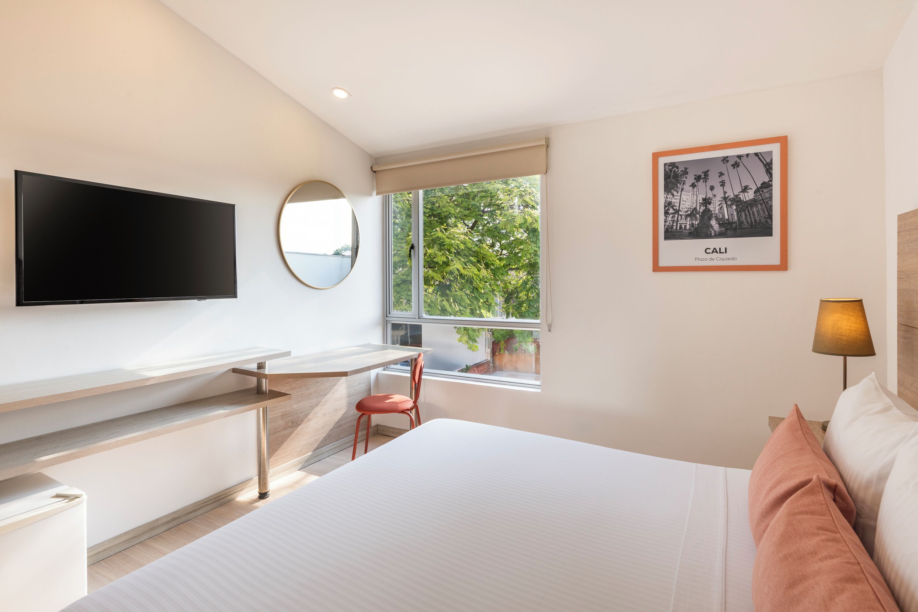 A modern hotel room with a bed, a desk, a chair, a TV, a window with a view of trees, and a framed picture on the wall.