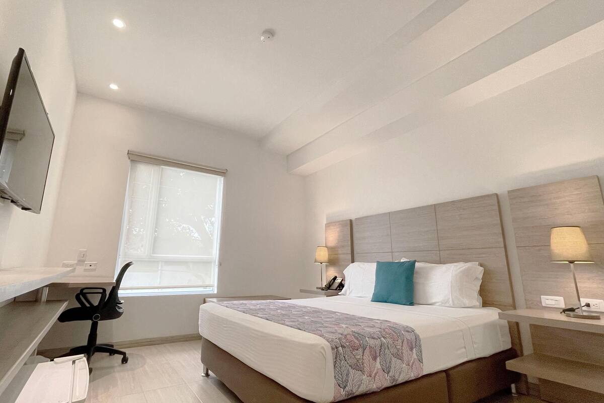 A hotel room with a bed, bedside tables, a desk with a chair, a TV mounted on the wall, and a window.
