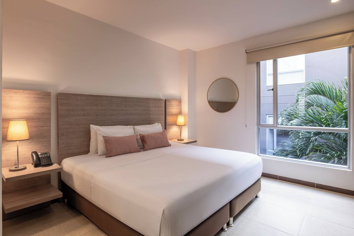 A hotel room with a large bed, bedside lamps, a round mirror, and a window with a view of greenery.