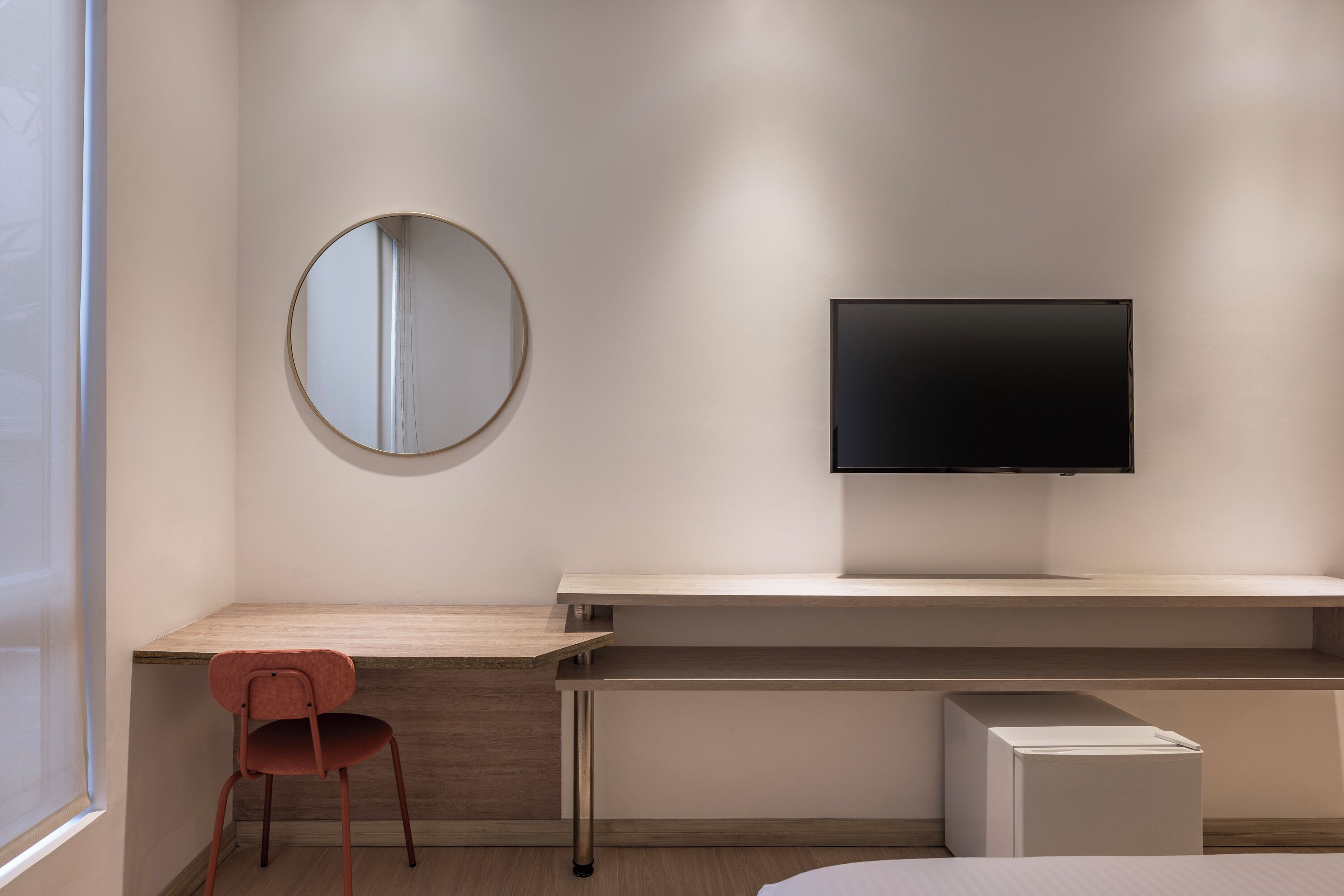 A modern hotel room with a wooden desk, a round mirror, a flat-screen TV, and a bed.