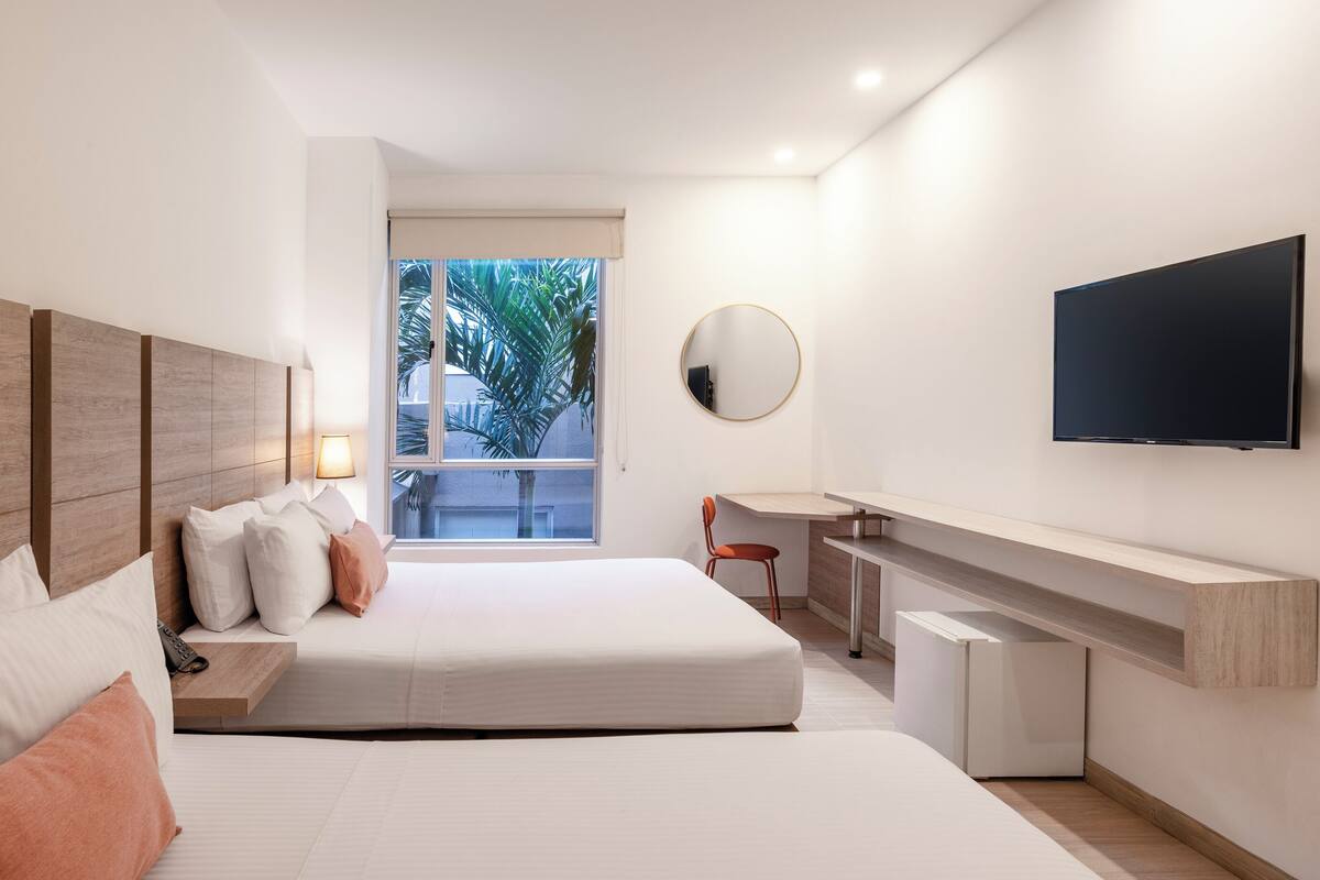 A modern hotel room with a large bed, a desk, a TV, and a window with a view of palm trees.