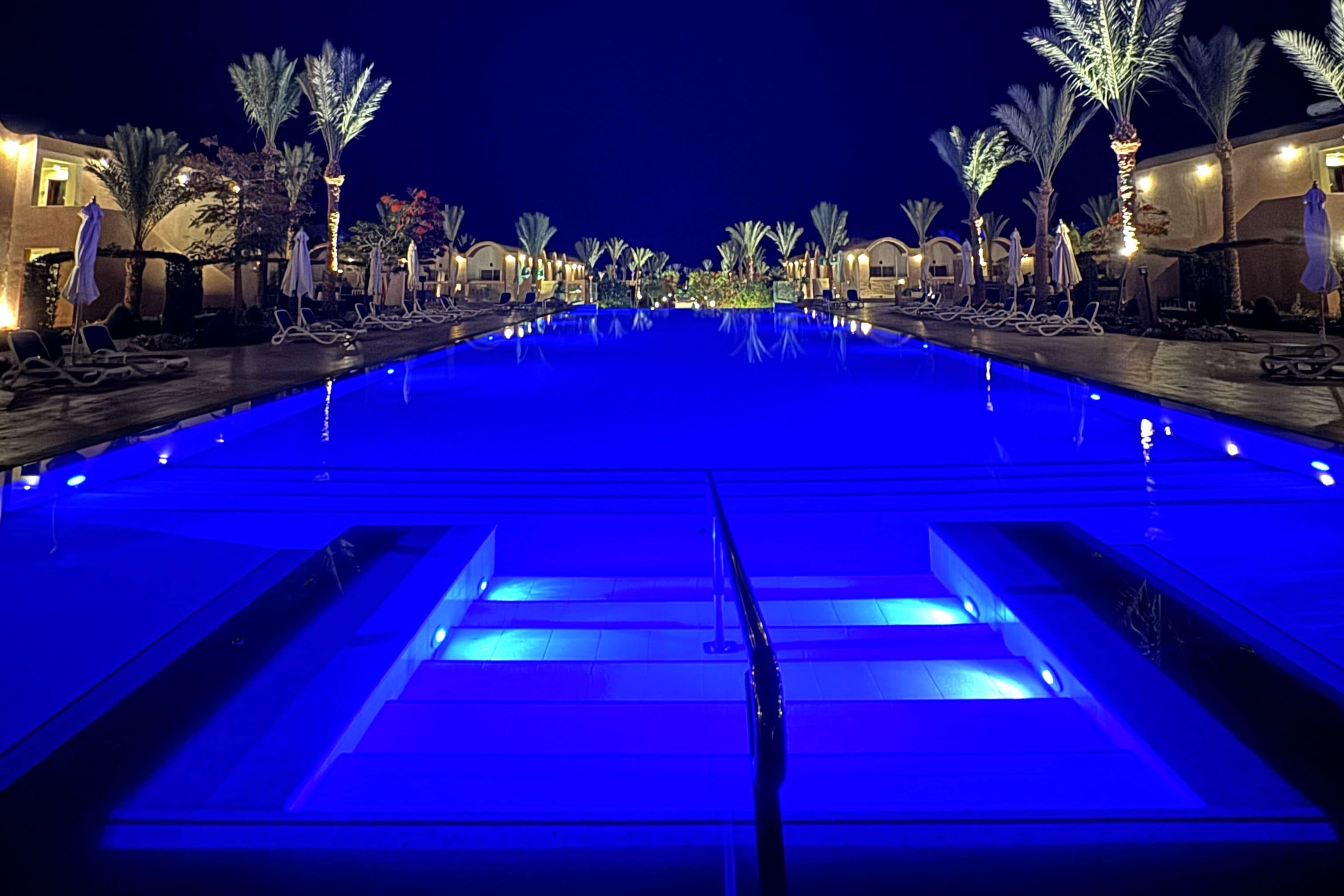 A pool with a blue-lit water feature, surrounded by palm trees and lounge chairs.
