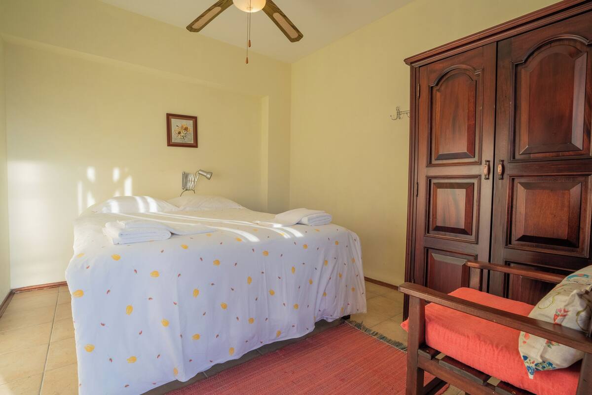 A bedroom with a bed, a wooden wardrobe, a chair, and a ceiling fan.