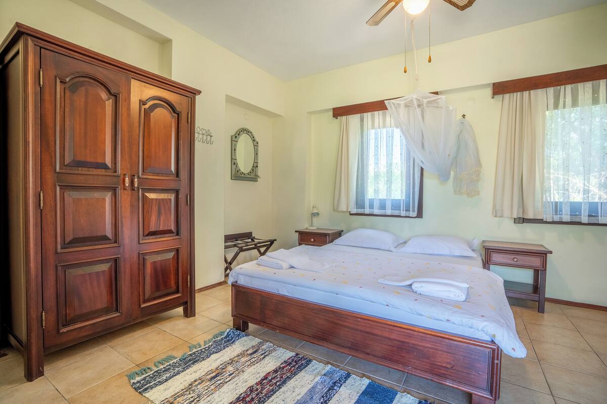 A bedroom with a wooden bed, a wardrobe, a bedside table, and a window with curtains.