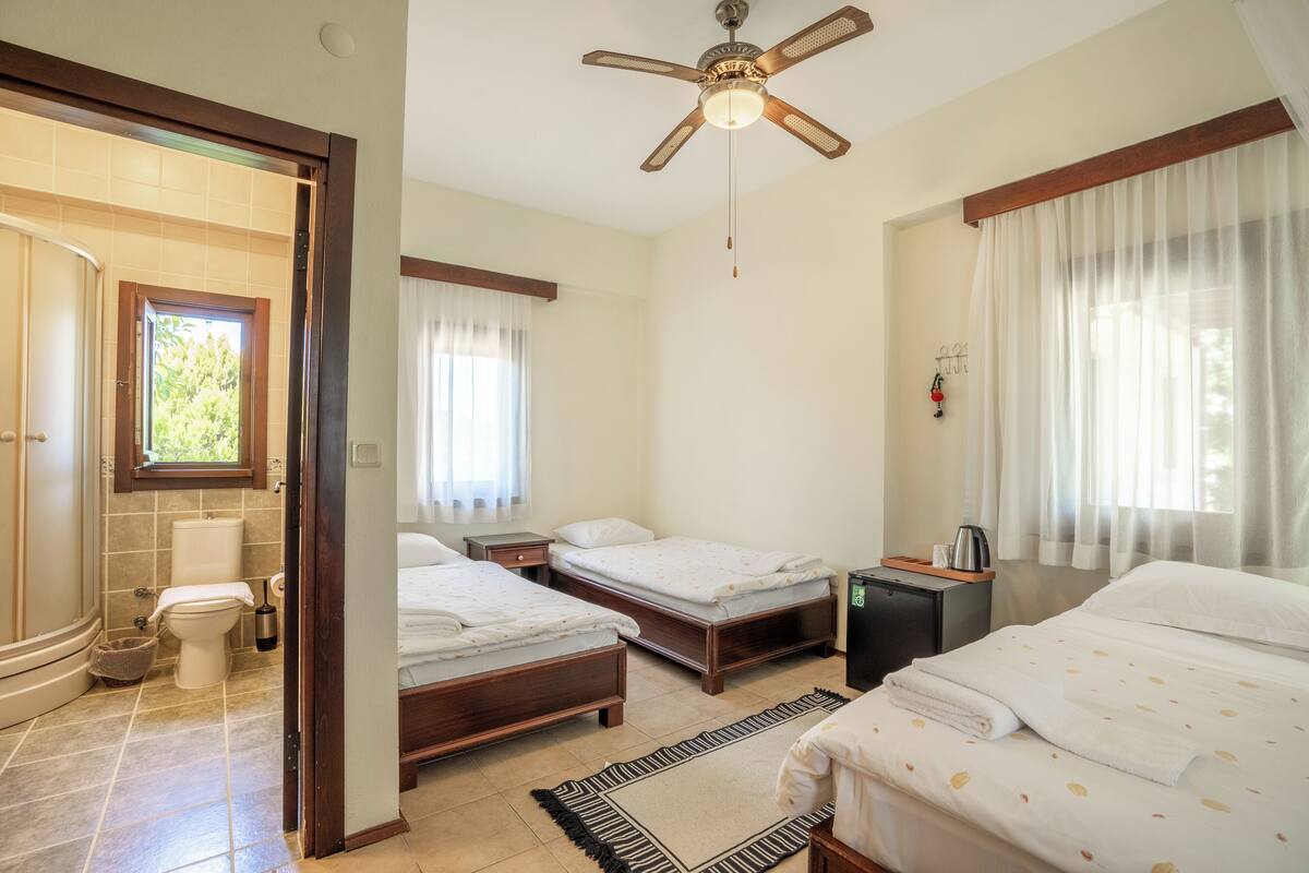 A hotel room with two beds, a ceiling fan, a bathroom with a toilet and sink, and a window with sheer curtains.