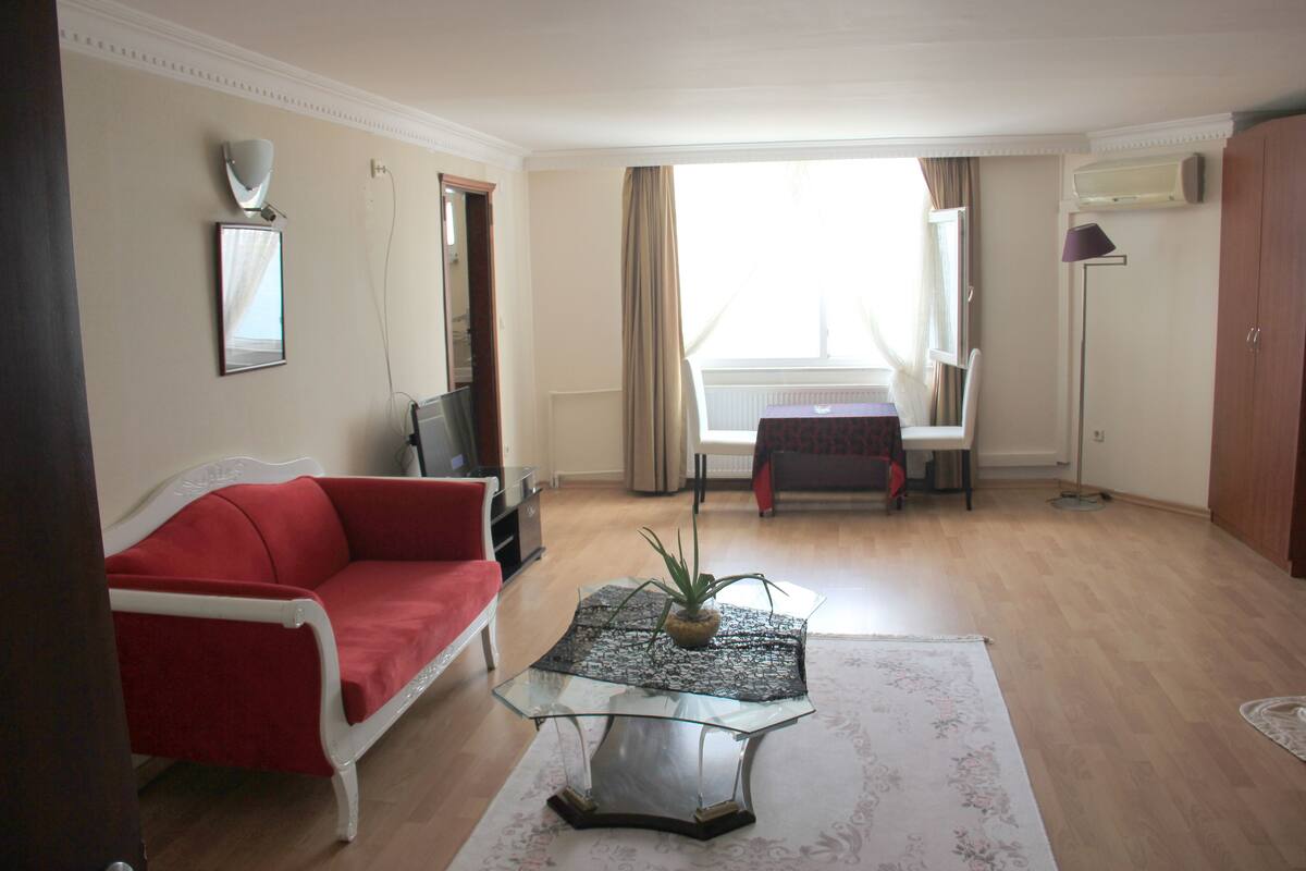 A living room with a red sofa, a glass coffee table with a plant, a white armchair, and a view of a balcony through a window.