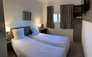 Habitación de hotel con dos camas, una mesita de noche con lámpara, televisión montada en la pared y ventana con cortinas.