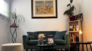 Ett vardagsrum med en grön soffa, ett soffbord med en vas med blommor, en inramad tavla på väggen, en bokhylla och en krukväxt.