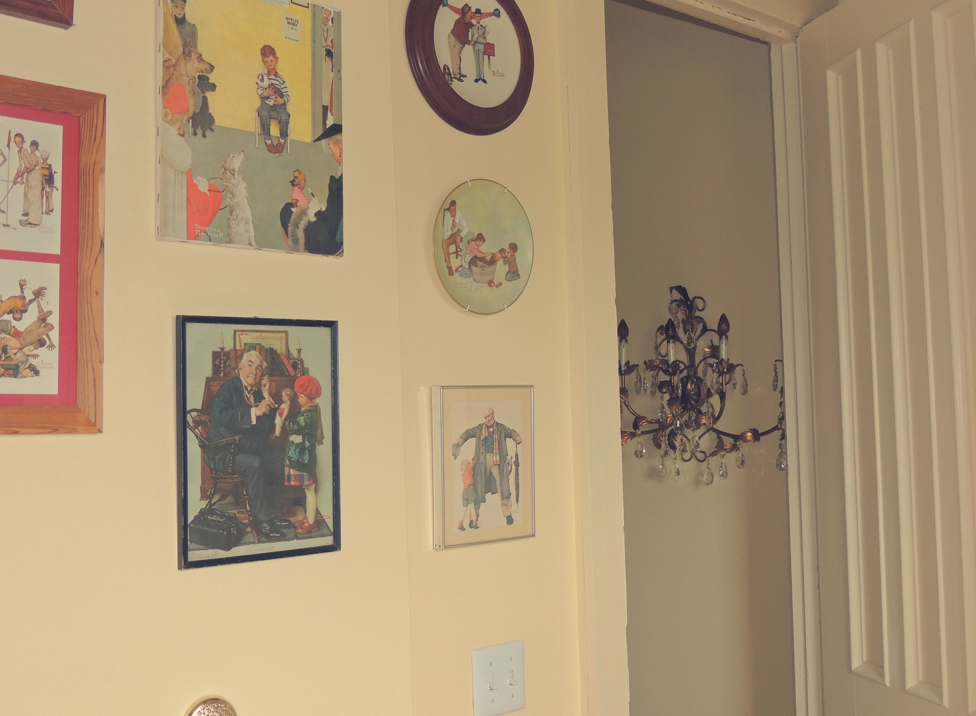 A room with various framed artworks on the walls, a chandelier, and a door leading to another room.