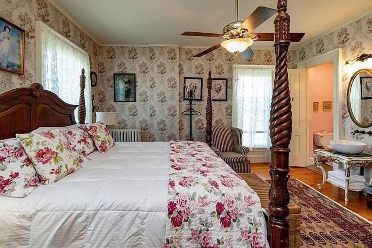 A bedroom with a four-poster bed, floral wallpaper, a ceiling fan, and a view of a bathroom with a sink and mirror.