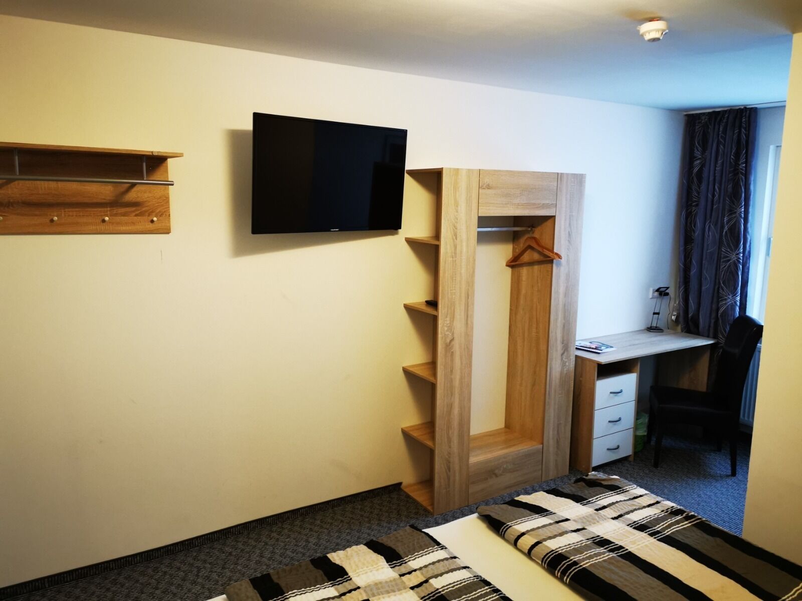 Quarto de hotel pequeno com uma cama, uma escrivaninha com cadeira, uma televisão na parede e um guarda-roupa com espelho.