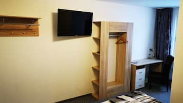 Quarto de hotel pequeno com uma cama, uma escrivaninha com cadeira, uma televisão na parede e um guarda-roupa com espelho.