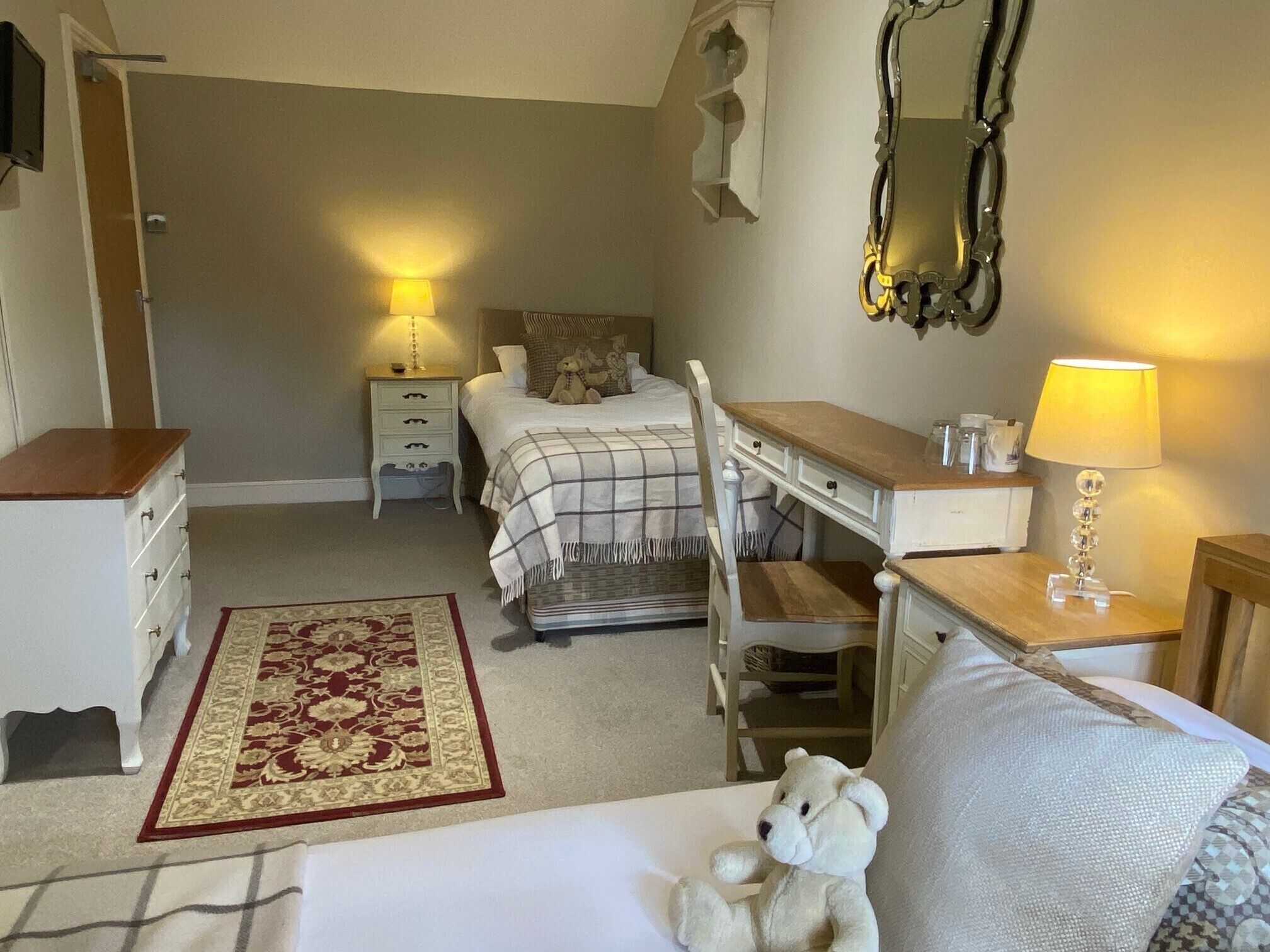 A bedroom with a bed, a desk, a chair, a nightstand, a lamp, a mirror, and a teddy bear.