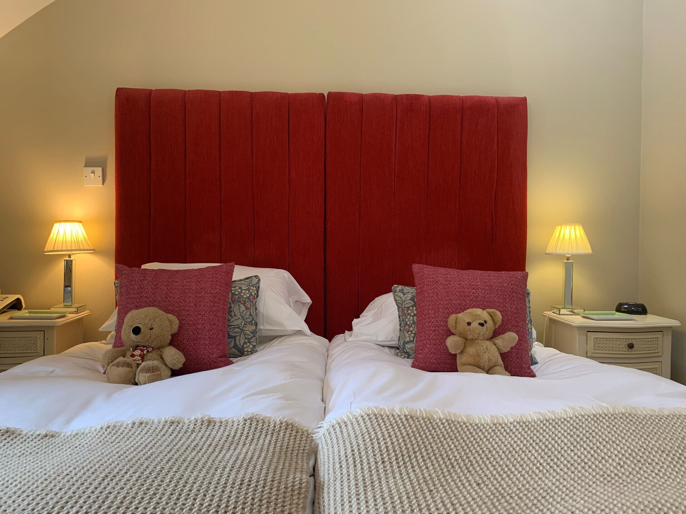 A double bed with a red headboard, two bedside lamps, and two teddy bears on pillows.