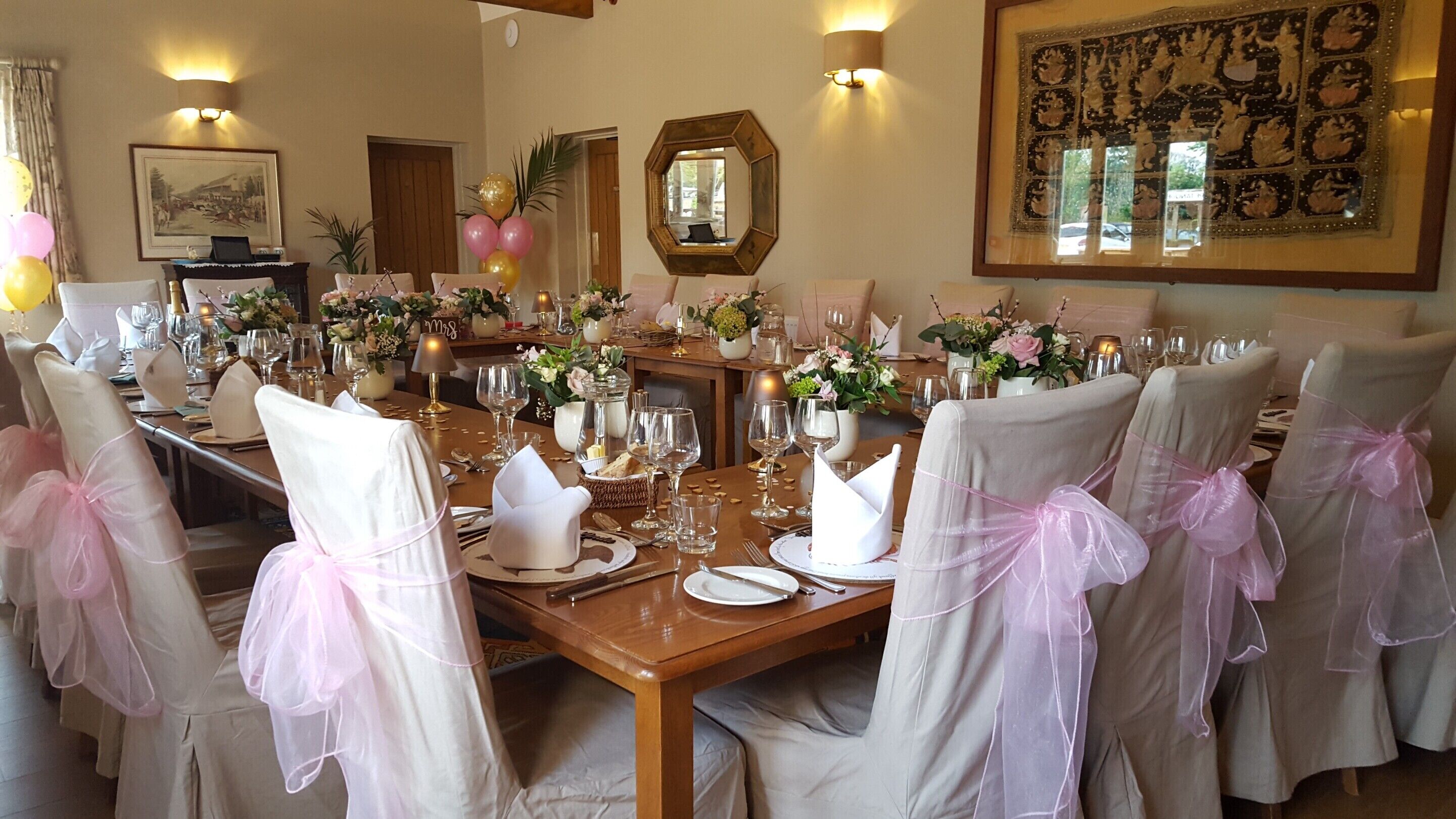 Un comedor formal con una larga mesa dispuesta para una comida, rodeada de sillas con lazos rosas y decorada con arreglos florales.