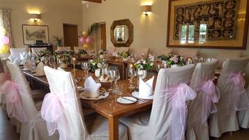 Un comedor formal con una larga mesa dispuesta para una comida, rodeada de sillas con lazos rosas y decorada con arreglos florales.