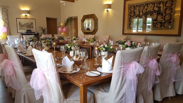 Un comedor formal con una larga mesa dispuesta para una comida, rodeada de sillas con lazos rosas y decorada con arreglos florales.