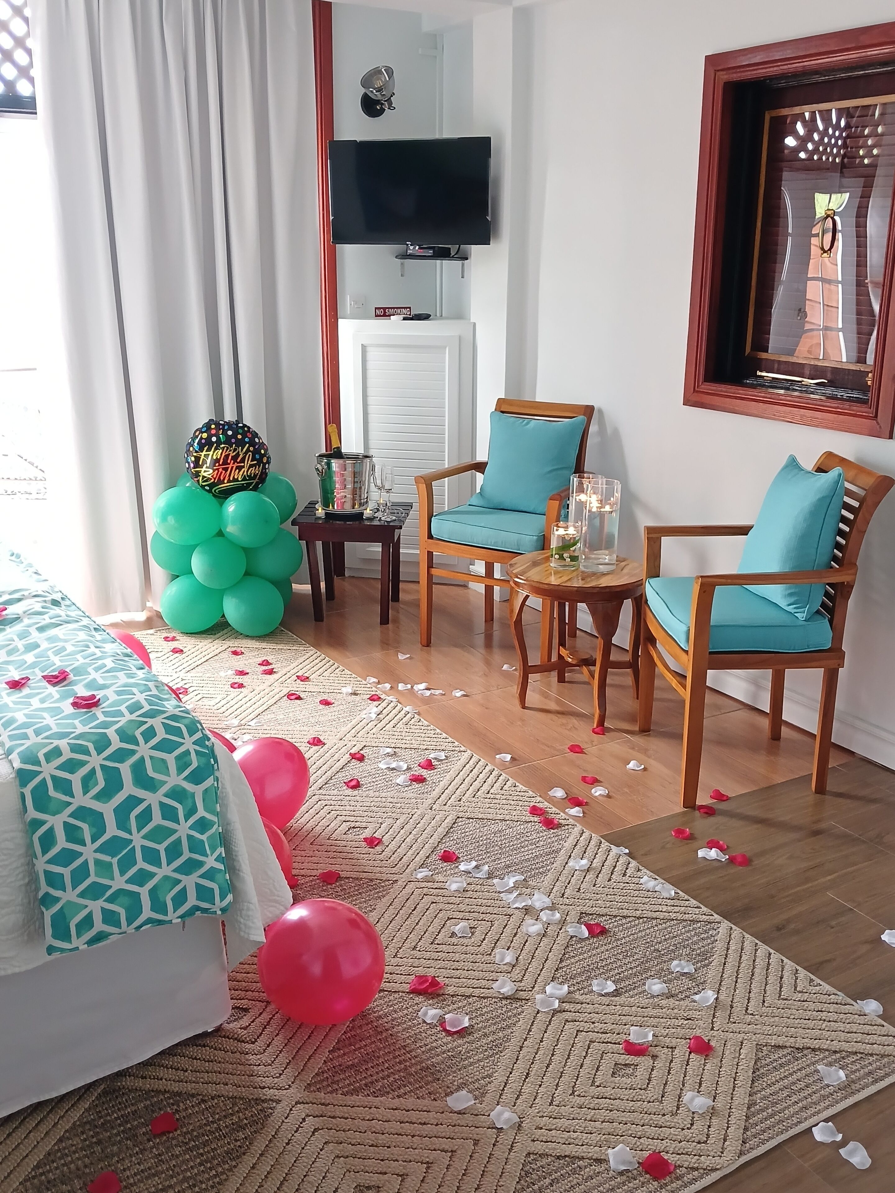 A hotel room with a bed, two chairs, a small table, a TV, and a decorative balloon arrangement.