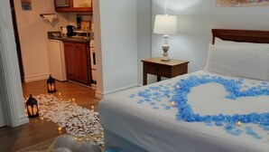 A hotel room with a bed adorned with blue rose petals, a nightstand with a lamp, and a painting on the wall. - Samfi Gardens (Soufrière)
