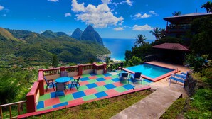 A pool area with a view of the ocean and mountains, surrounded by greenery and a red-roofed building. - Samfi Gardens (Soufrière)