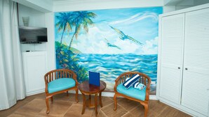 A room with a mural of a beach scene, two wooden chairs with cushions, a small round table, and a white wardrobe. - Samfi Gardens (Soufrière)