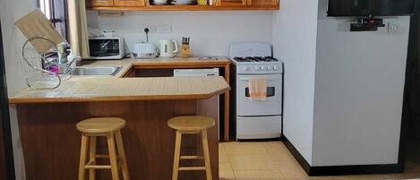 Une cuisine compacte avec des meubles en bois, un four à micro-ondes, une cuisinière et un téléviseur. La pièce comprend deux tabourets et un lit.