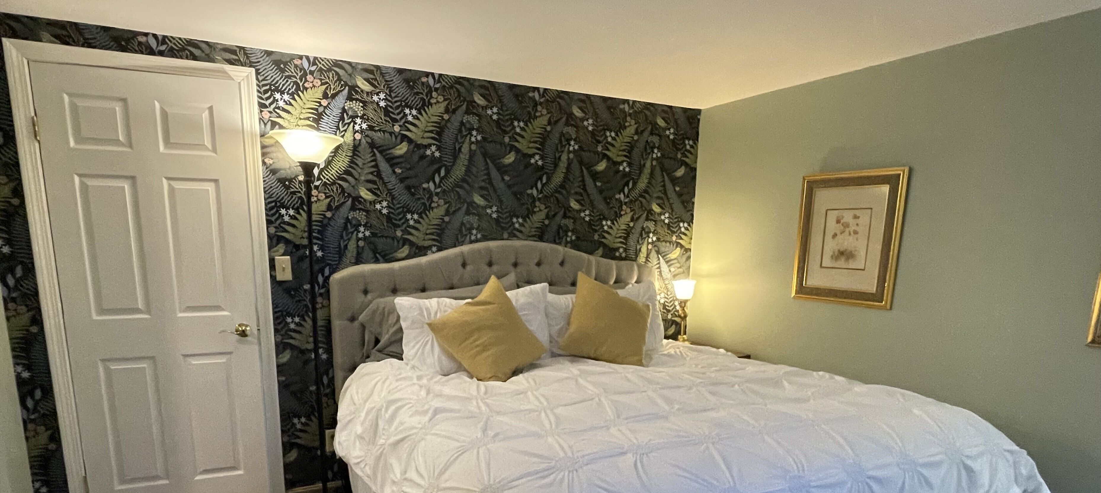 A bedroom with a bed, a door, a wall with a floral pattern, and a framed picture.