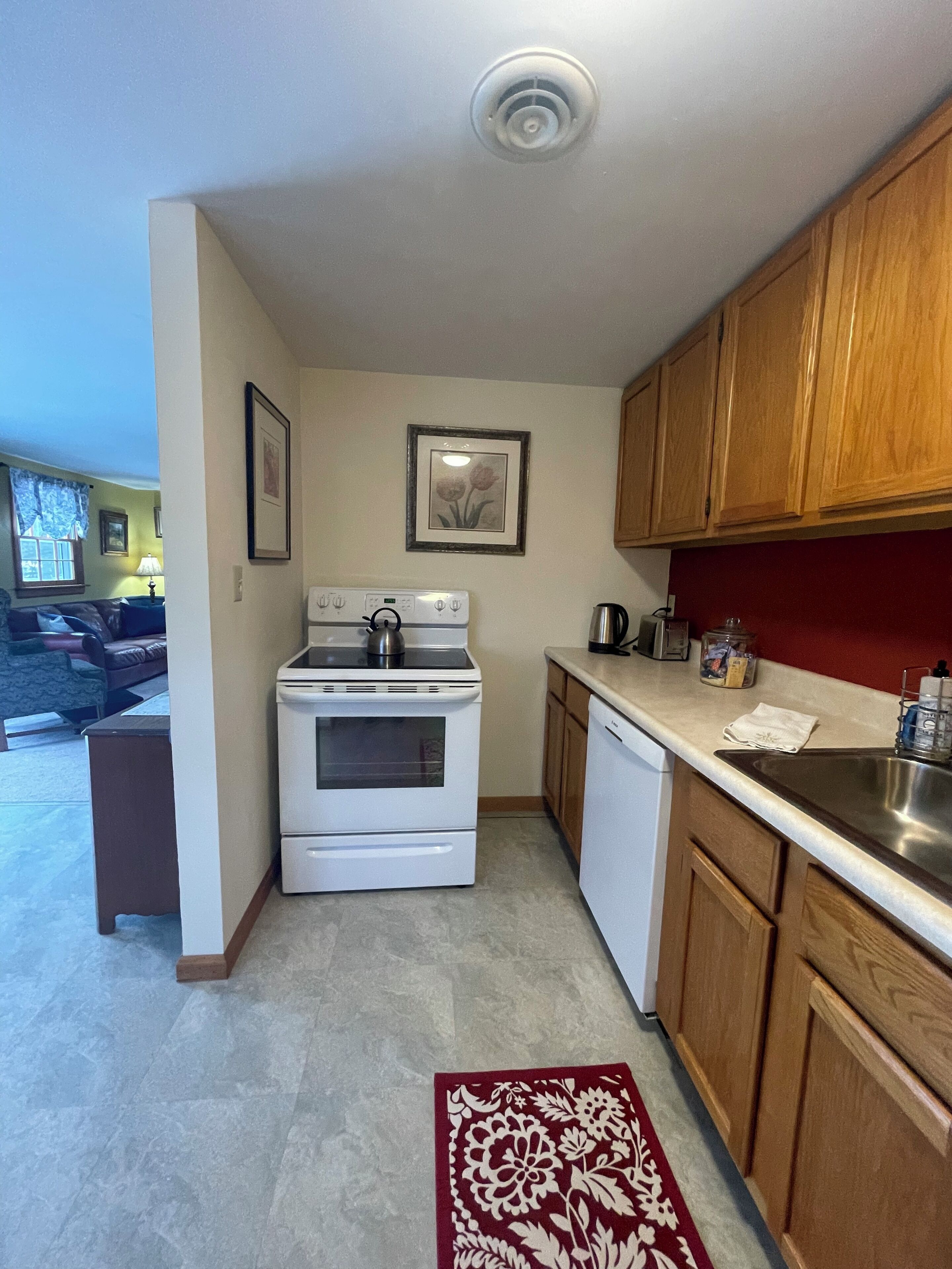 A compact kitchen with a stove, oven, and sink, wooden cabinets, and a red rug on the floor.