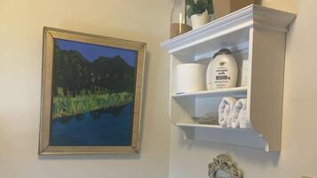 A bathroom with a toilet, a framed painting, a shelf with toiletries, and a small plant on the toilet.