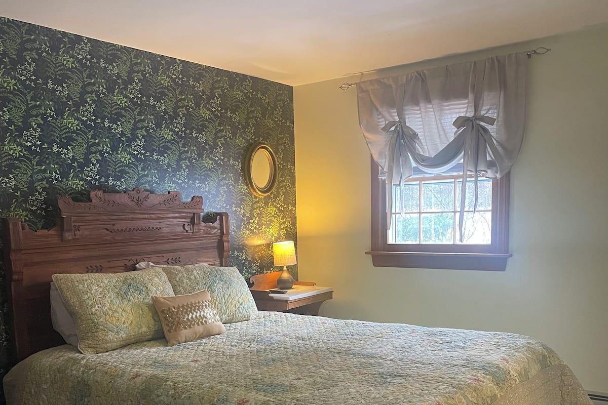 A bedroom with a large bed, a wooden headboard, a nightstand with a lamp, a window with curtains, and a wall with a floral wallpaper.