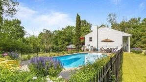 Una piscina rodeada de abundante vegetación y una casa blanca con un patio cercado.