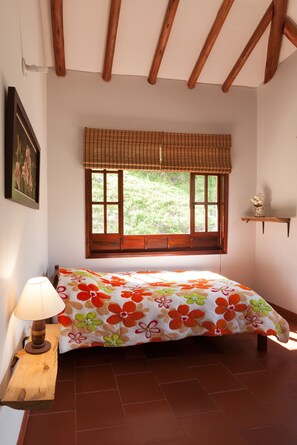 Quarto com cama, janela com persiana xadrez, luminária de cabeceira e uma pequena estante com flores.