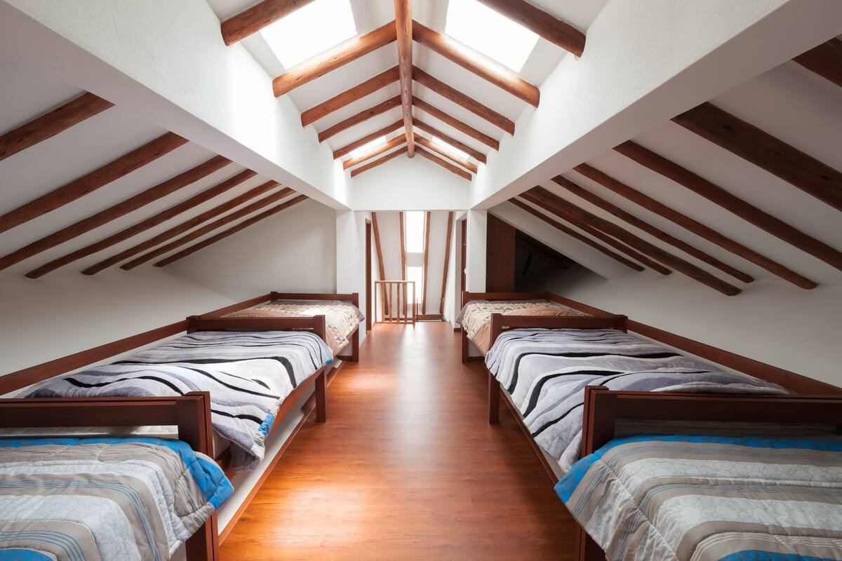 Una habitación con dos camas de madera alineadas a lo largo de un pasillo central, techo inclinado con vigas de madera y un tragaluz.