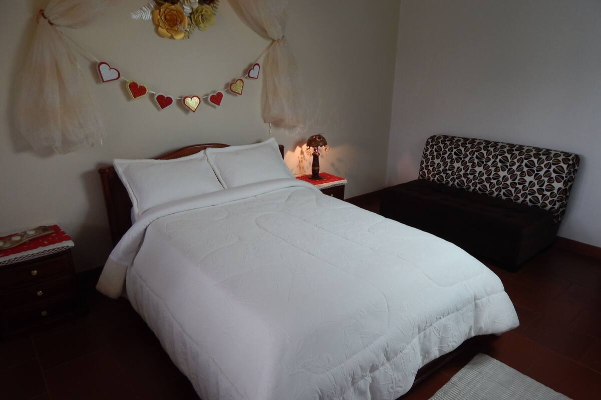 Una cama bien hecha con un edredón blanco, una mesita de noche con una lámpara y un banco estampado.