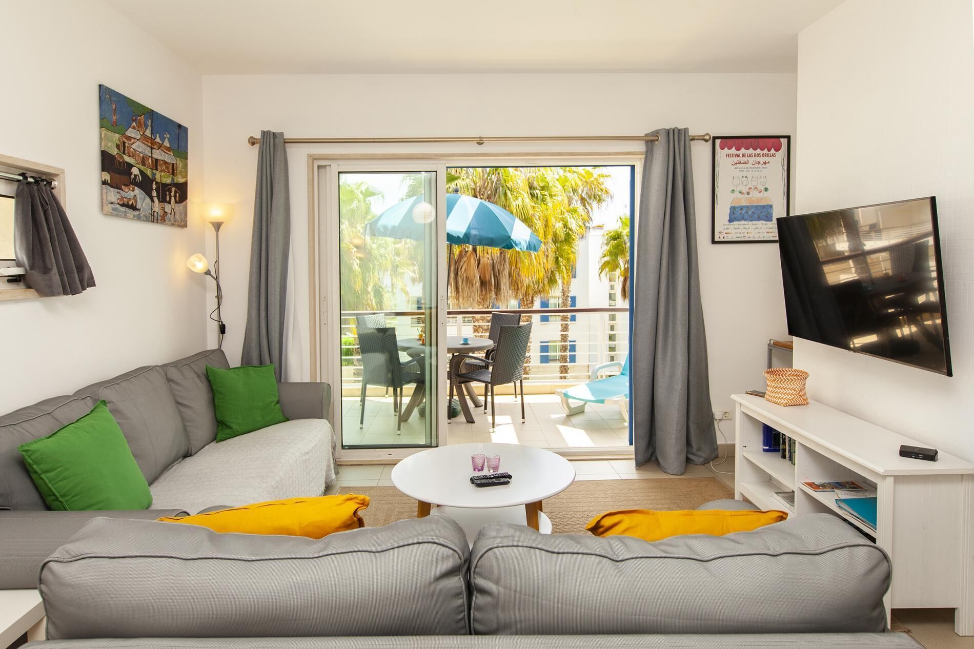 A modern living room with a grey sofa, a small white table, a TV, and a balcony with outdoor furniture.