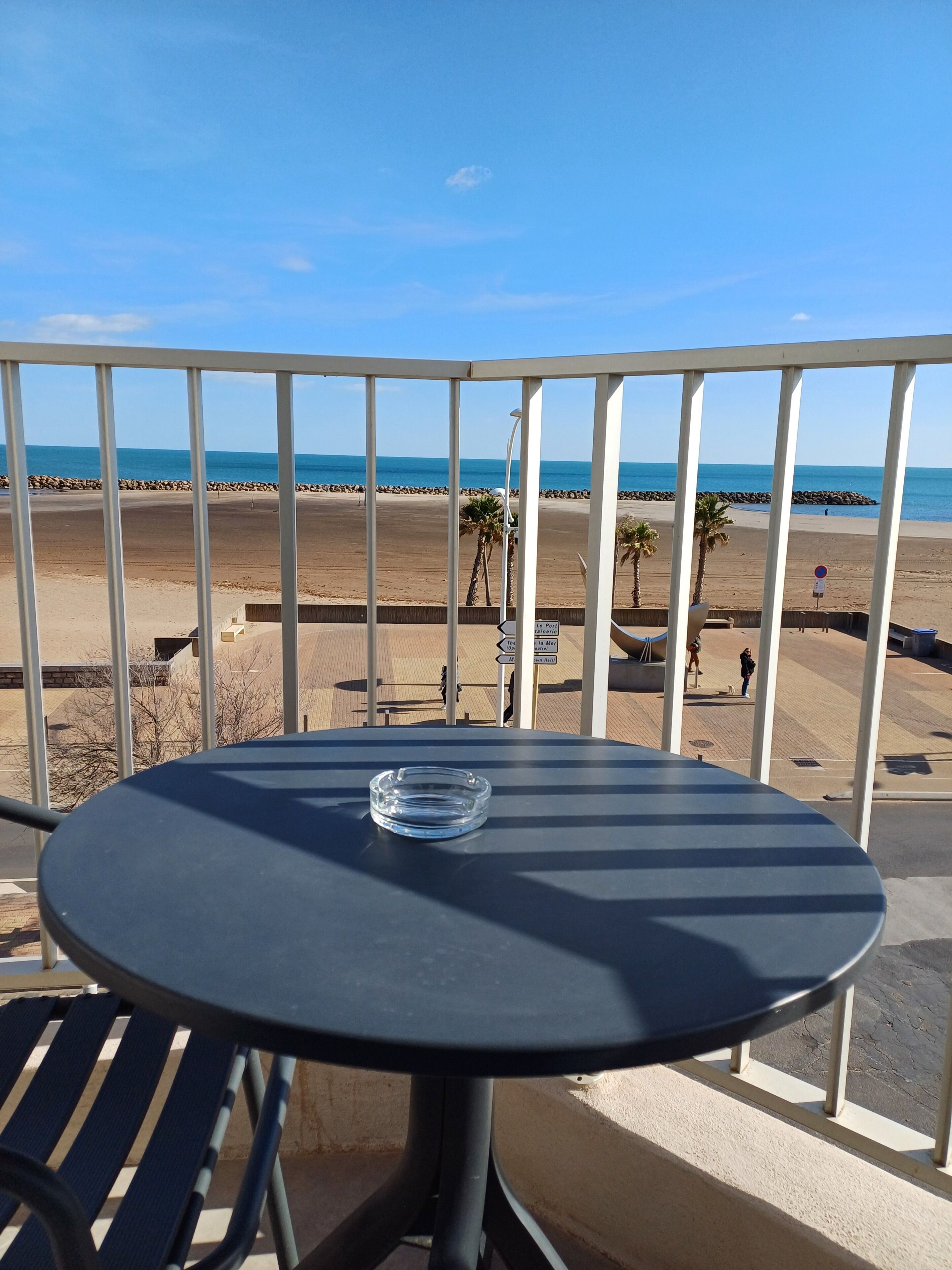 Un balcón con mesa y sillas, con vista a una playa y cielo despejado.
