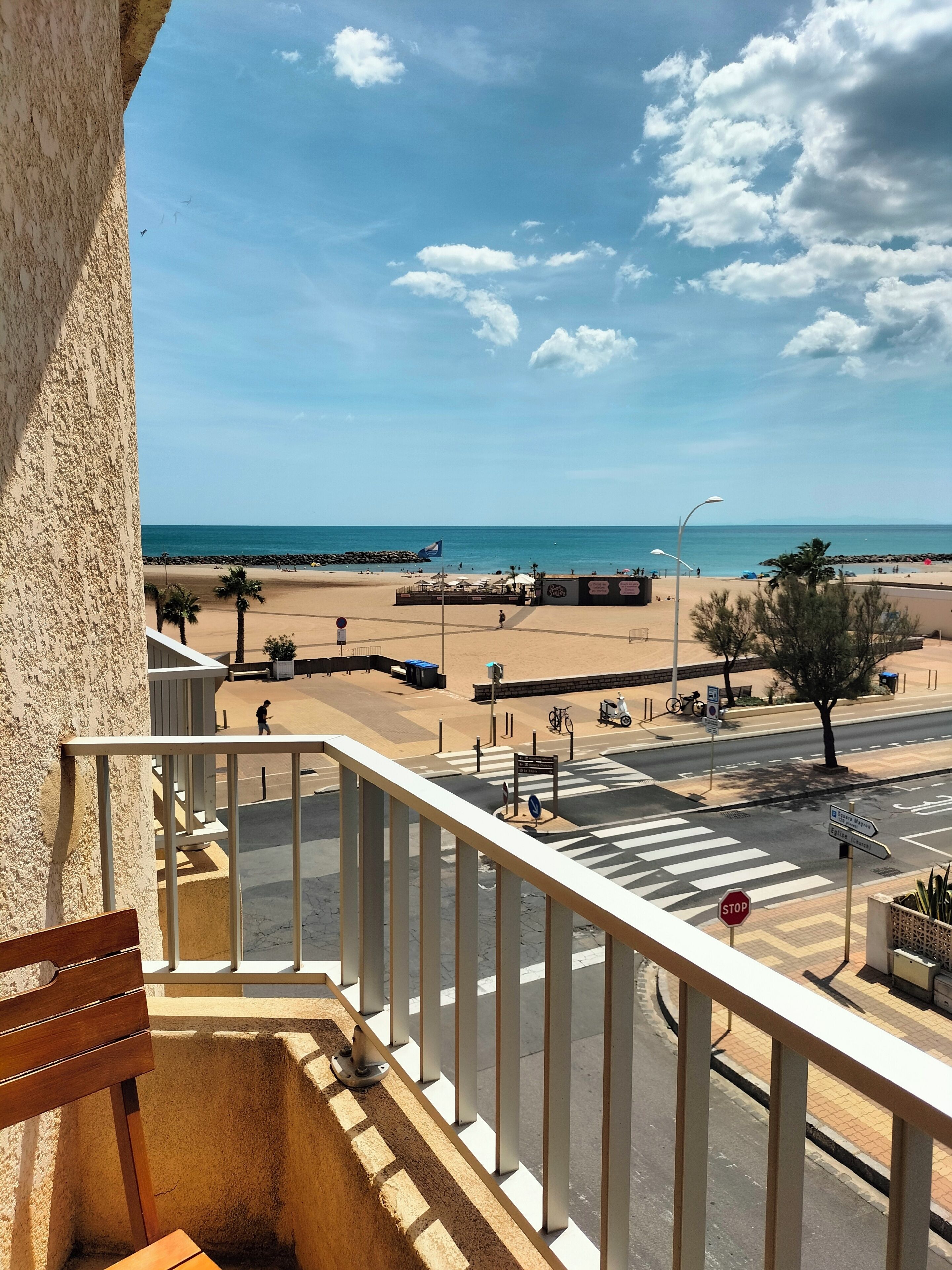Un balcón con vista a una playa, cielo despejado, un paso de peatones y una calle con vehículos.