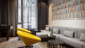 Un salon moderne avec un fauteuil jaune, un canapé gris, une petite table et un mur décoré de rubans colorés.