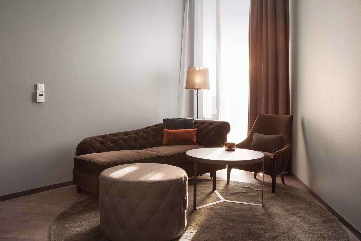 A modern hotel room with a sectional sofa, a round coffee table, and a tufted ottoman.